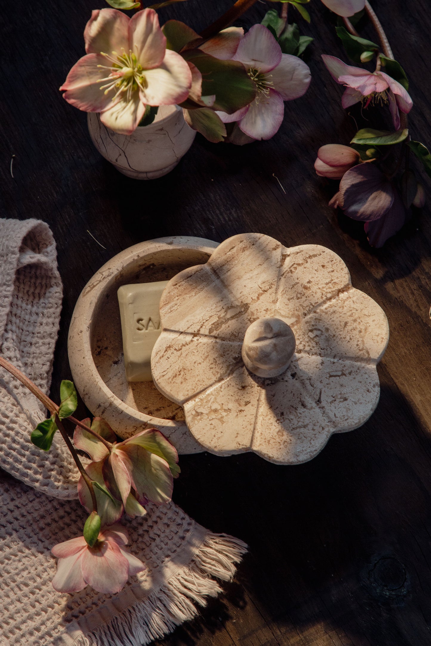 A white travertine trinket box with a floral design, displayed on a wooden surface alongside pink flowers and a beige cloth.